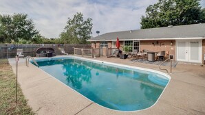 Une piscine rectangulaire dotée d’une terrasse en bois et de chaises longues, d’un espace barbecue et d’un patio couvert aménagé avec du mobilier d’extérieur.
