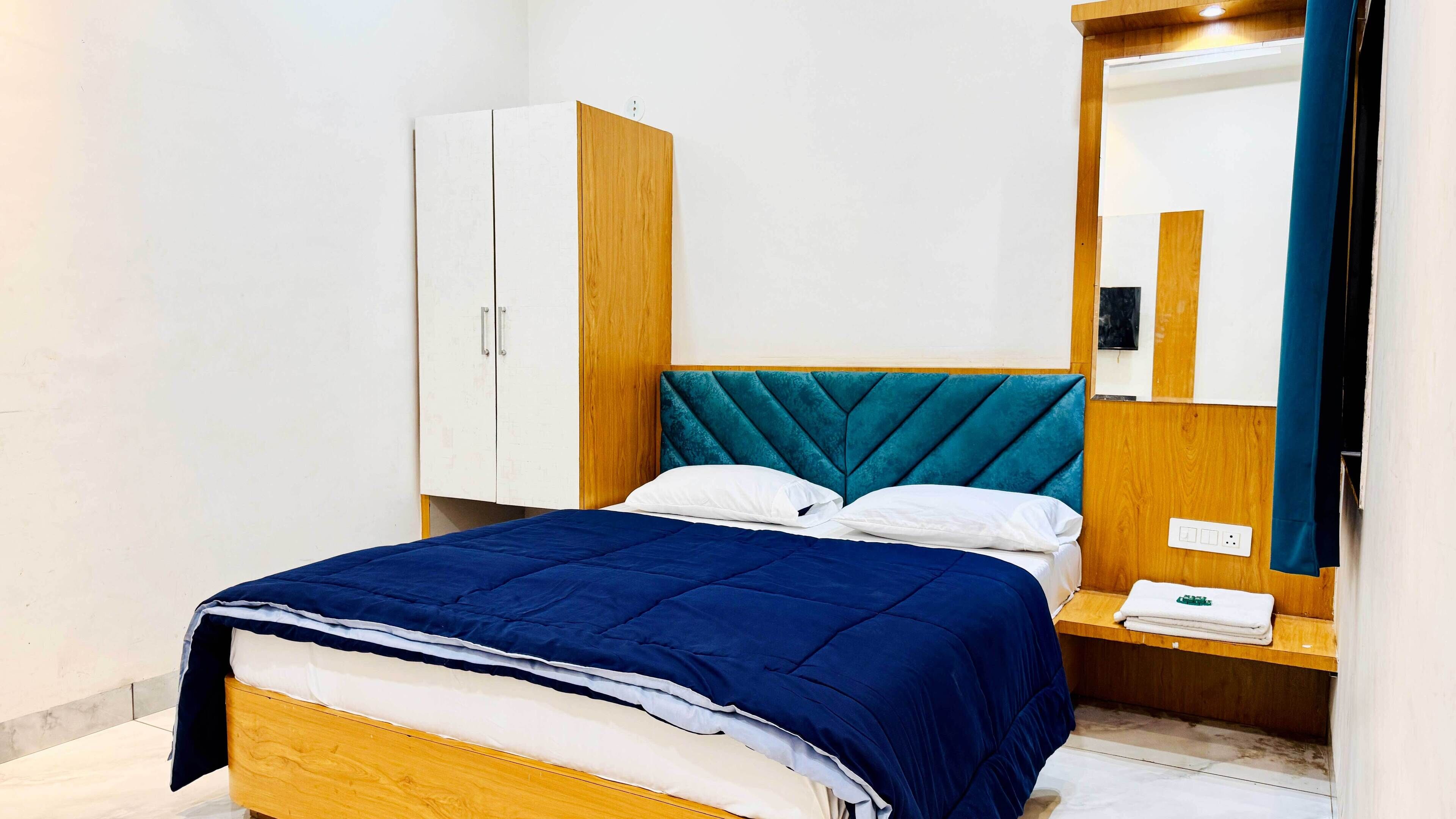 A neatly made bed with a blue bedspread, white pillows, and a teal headboard. There is a wooden nightstand with a folded towel and a mirror above it. A white wardrobe is visible to the left.