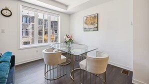 Een moderne eetruimte met een glazen tafel en twee stoelen, een groot raam met uitzicht op gebouwen, een klok aan de muur en een ingelijste foto.