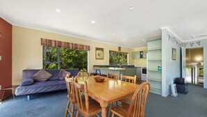 A living room with a sofa, wooden dining table, and chairs. There is a kitchen area with a counter and cabinets. - Kereru Retreat at Moetapu Bay (Moetapu Bay)