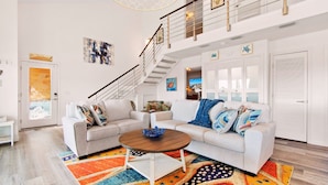 Un salon spacieux avec un canapé blanc, une table basse et un tapis coloré. Un escalier avec une rampe mène à l’étage supérieur.