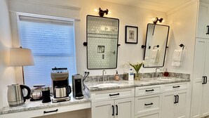 Eine moderne Küche mit weißen Schränken, einer Marmorarbeitsplatte, einer Kaffeemaschine, einem Wasserkocher und einem Waschbecken mit Spiegel darüber.