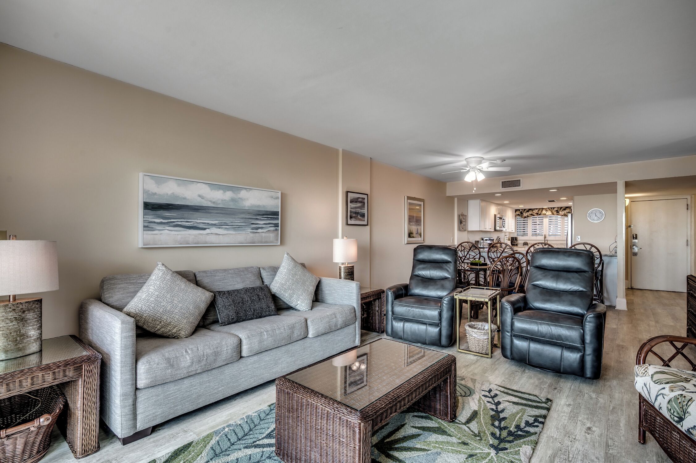 A living room with a grey sofa, two black leather chairs, a glass coffee table, and a large painting of the ocean.