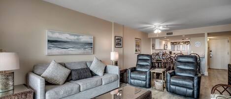 A living room with a grey sofa, two black leather chairs, a glass coffee table, and a large painting of the ocean.