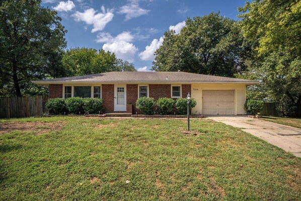 A single-story brick house with a front garage, surrounded by a well-maintained lawn and trees.