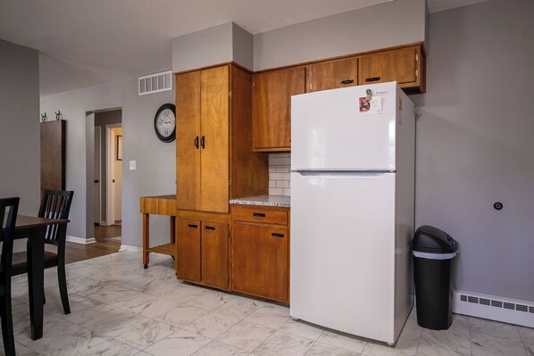 A kitchen with wooden cabinets, a white refrigerator, a clock, and a trash bin.