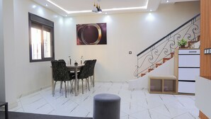 Un comedor con una mesa y sillas, un reposapiés gris, una escalera con barandilla y una obra de arte colgada en la pared.