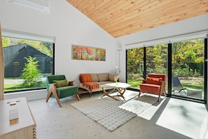 Una sala de estar moderna con techo de madera, ventanas amplias, un sofá, sillas y un cuadro.