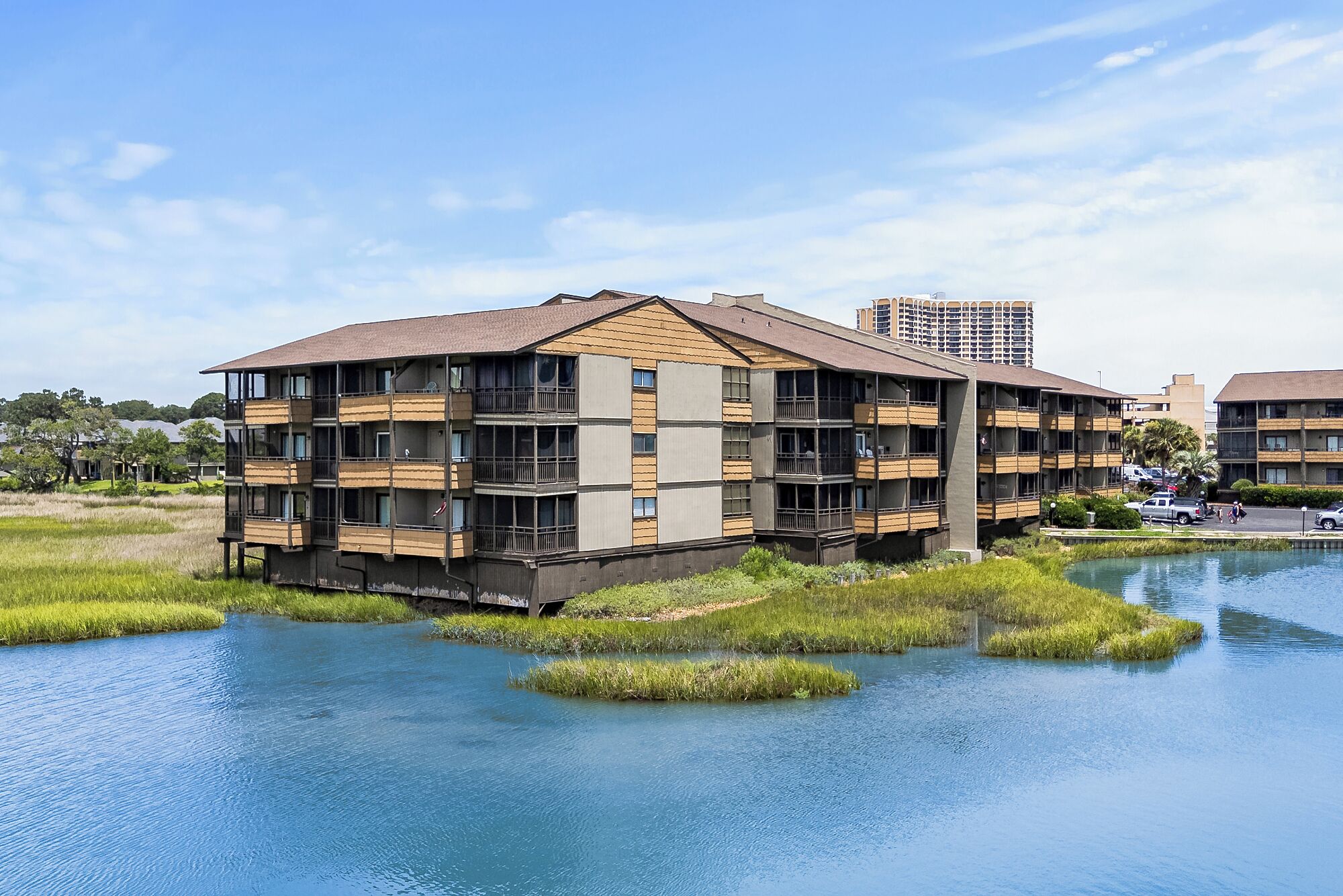 A multi-story building with balconies, situated by a body of water with grassy banks.