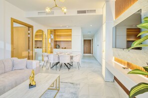 A modern living room with a sofa, coffee table, and dining area. There is a large window on the right side. - PVH - Canal Frontage 1BR - Millennium (Dubai)