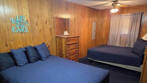 Ein Zimmer mit Holzwänden, einem Deckenventilator, einem Bett mit blauer Bettdecke, einem Schminktisch und einem Spiegel.
