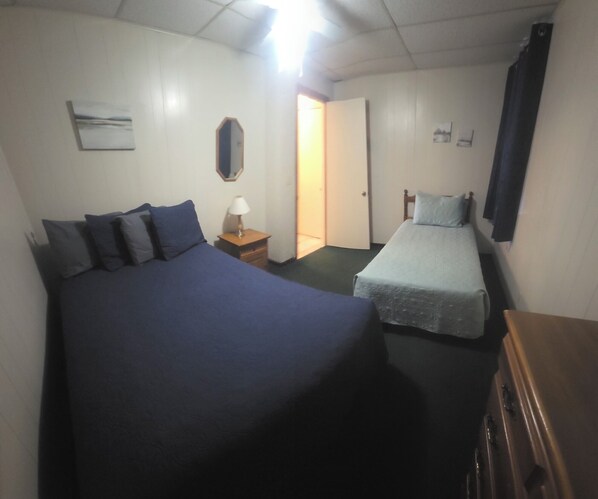 Une pièce comprenant un grand lit, un plus petit lit, une table de chevet avec une lampe, une commode et une porte donnant sur une autre pièce.