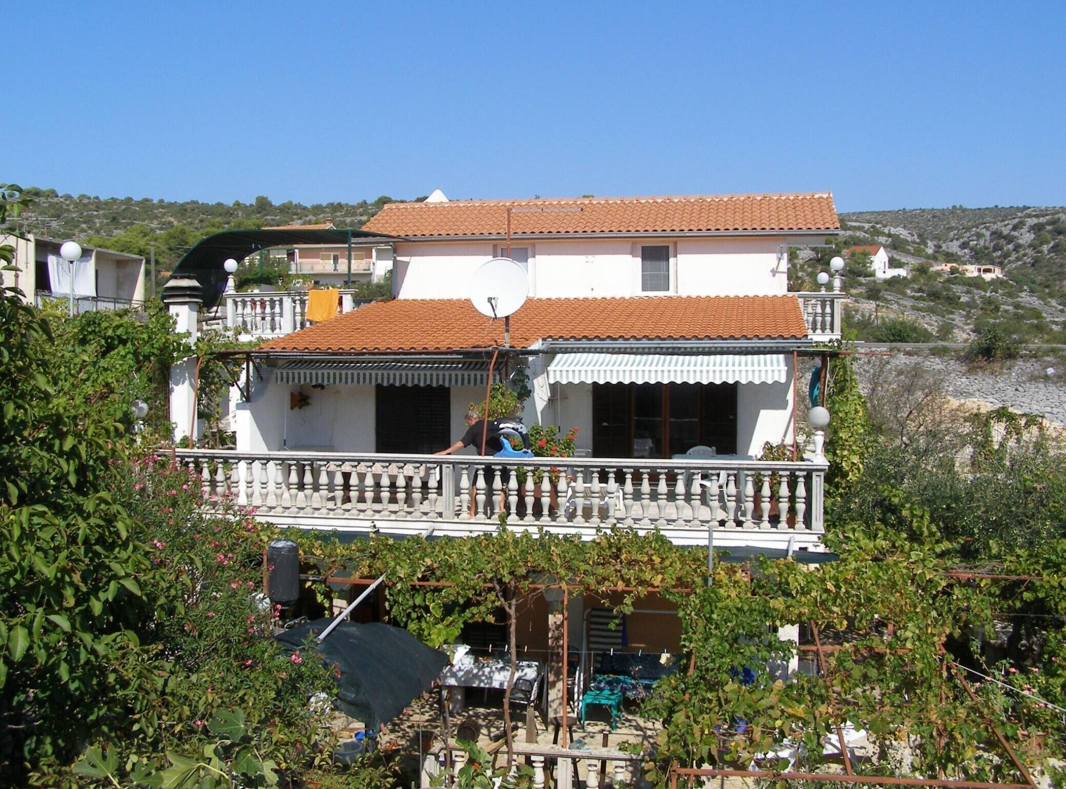 A two-story house with a balcony, surrounded by greenery and a hillside in the background.