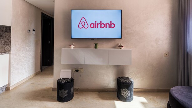 Un salon moderne avec un téléviseur fixé au mur qui affiche le logo d’Airbnb, une console blanche en dessous et deux tabourets noirs décoratifs.