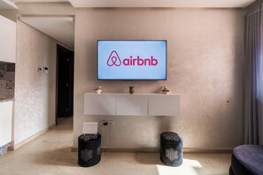 A modern living room with a mounted television displaying the Airbnb logo, a white console table below, and two decorative black stools. - Charming 2-bedroom apartment with AC in nice Marrakech (Marrakech)