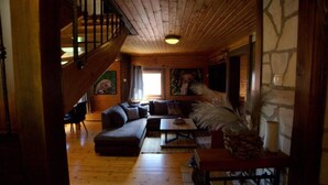 A cozy cabin interior with a wooden staircase, a large sofa, a dining table, and a stone wall. - Brezova Poljana Lodge with Sauna and Hot-Tub - Five Bedroom Holiday Home (Vrbovsko)