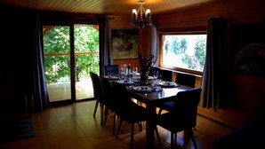 A dining area with a wooden table set for four, surrounded by chairs. A chandelier hangs from the ceiling, and there is a large window providing a view of greenery outside. - Brezova Poljana Lodge with Sauna and Hot-Tub - Five Bedroom Holiday Home (Vrbovsko)