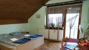 Una habitación acogedora con techo de madera, una cama con manta azul, una mesa con mantel rojo y fruta, y una ventana con cortinas.