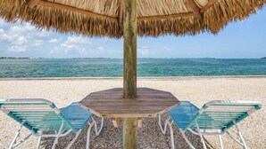 A wooden table with a thatched umbrella and two green metal chairs on a pebble beach. - Waterfront 2BR, Dock,Golf Cart, trailer parking,Open Water Views Venture Out 320 (Cudjoe Key)
