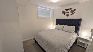 Une chambre à coucher avec une tête de lit capitonnée, un lit simple avec une literie à motifs, une table de chevet avec une lampe, une petite fenêtre avec des stores et un élément décoratif fixé au mur.