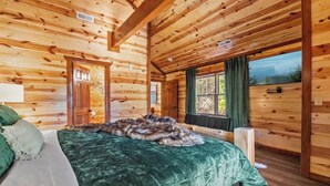 A log cabin bedroom with a bed, a window with curtains, and a view of the outdoors. - Amazing Luxury NEW 8bdrm Cabin-Pool-Arcades-Gazebo (Sevierville)