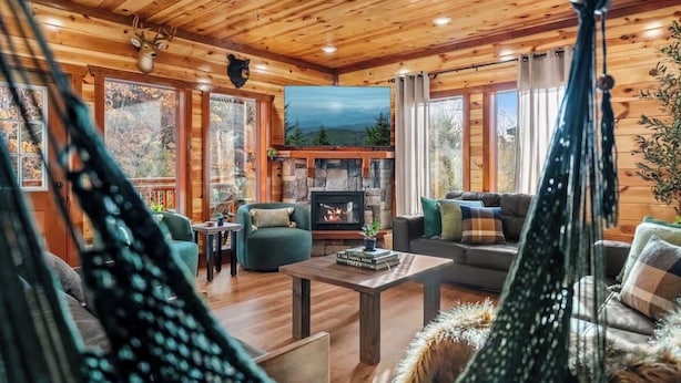 A cozy cabin interior with a stone fireplace, wooden walls, and a view of the forest through large windows. - Amazing Luxury NEW 8bdrm Cabin-Pool-Arcades-Gazebo (Sevierville)
