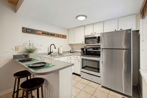 A modern kitchen with stainless steel appliances, a central island with bar stools, and a decorative wooden sign on the wall. - 1BD King Condo – Walking Distance to PCMR on Park Ave (Park City)