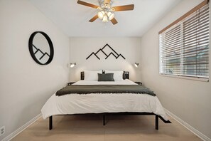 Ein Schlafzimmer mit einem Bett, einem Deckenventilator, einem wandmontierten Spiegel und einem Fenster mit Jalousien.