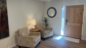A room with two patterned armchairs, a small table with a lamp, a round mirror, and a wooden door.