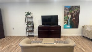 A living room with a beige sofa, a dark wooden entertainment unit with a flat-screen TV, a plant on a metal shelf, and a large waterfall painting on the wall.