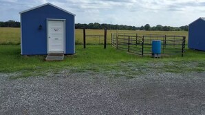 A blue building with a white door, a metal gate, and a blue barrel in a grassy field. - Friesian Cabin (Heavener)