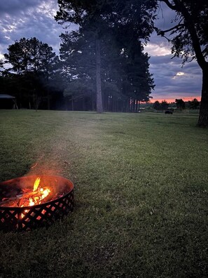 A grassy outdoor area with a fire pit in the foreground and trees in the background.