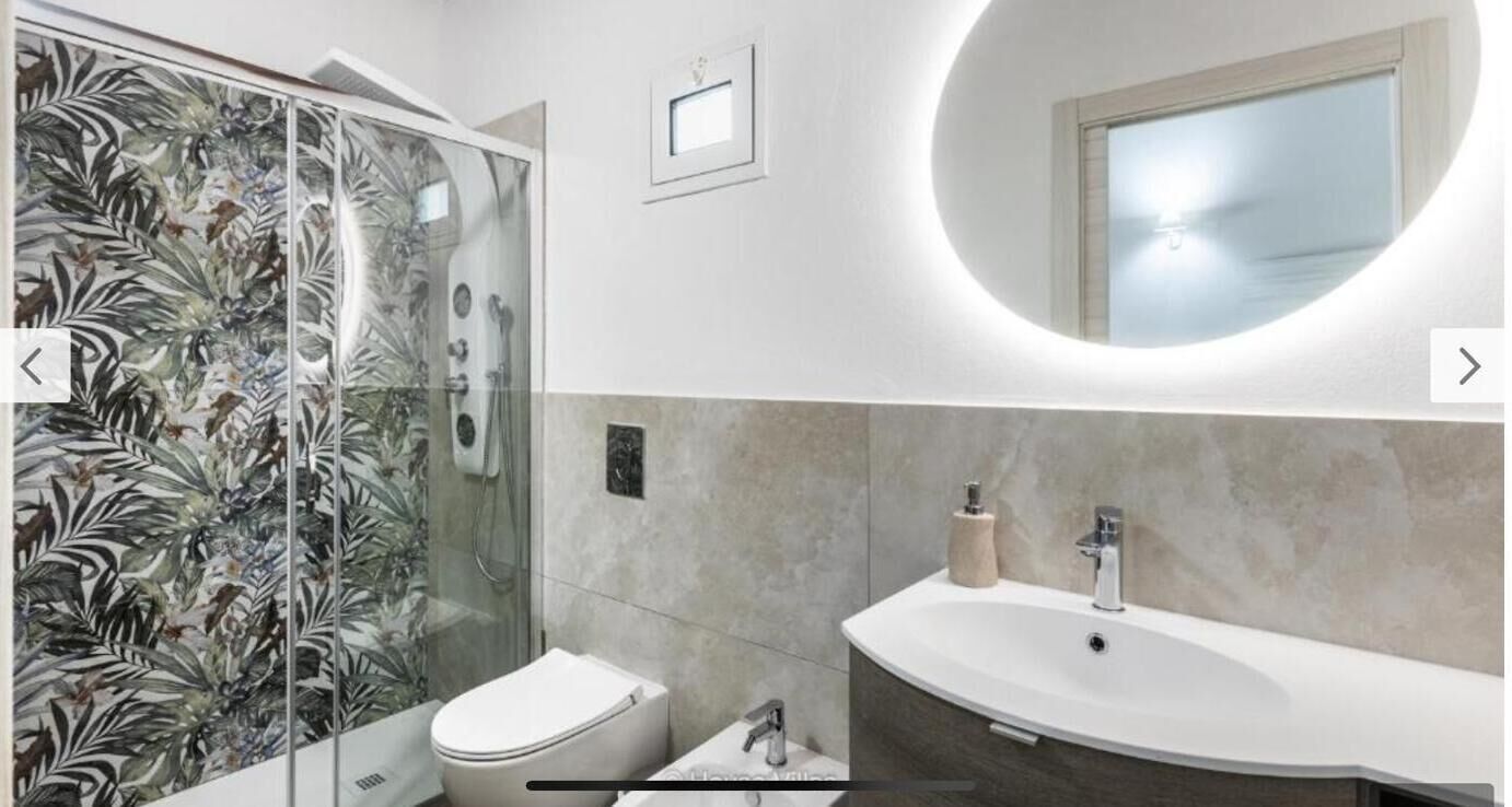 A bathroom with a glass shower enclosure, a toilet, a sink with a mirror, and a wall-mounted light fixture.