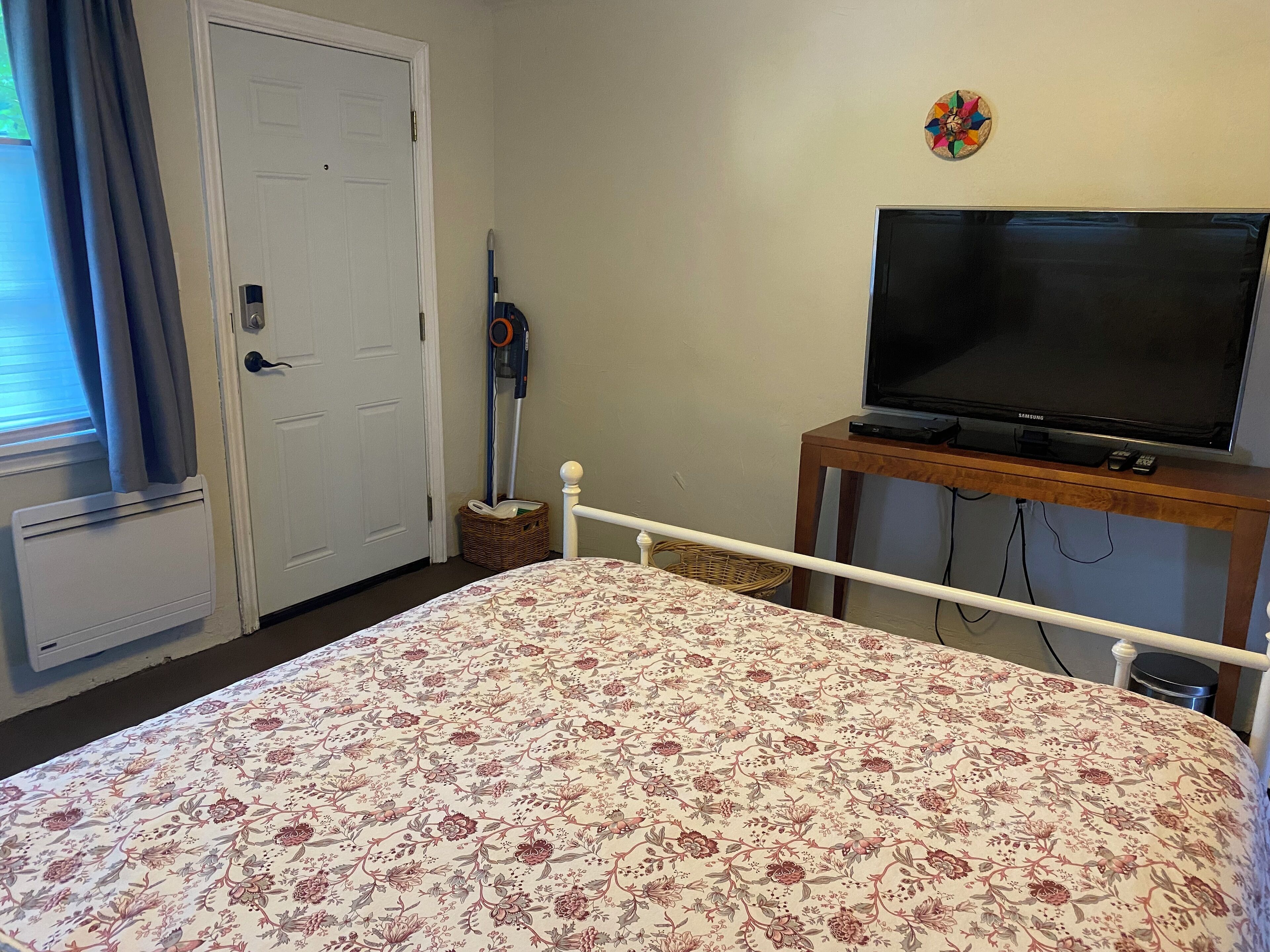 Un dormitorio con una cama, un televisor sobre un soporte de madera, una puerta y una ventana con cortinas.