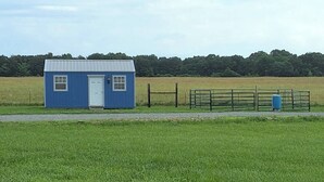 A small blue building with a white door, situated in a grassy field with a metal fence and a blue barrel nearby. - Morgan Cabin (Heavener)