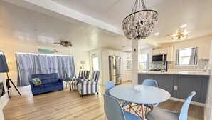 Een moderne woonkamer met een blauwe bank, gestreepte fauteuils en een ronde tafel met blauwe stoelen. Een kroonluchter hangt aan het plafond, en in de achtergrond is een keuken zichtbaar met witte kasten en een betegelde spatwand.