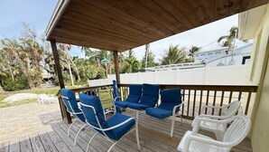 Een overdekt terras buiten met blauwe en witte lounge stoelen, een kleine tafel en een houten balustrade.