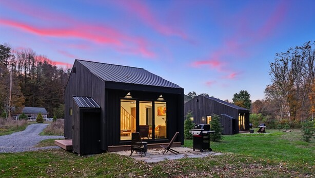 Una cabana moderna de color fosc, amb una gran porta de vidre i finestres, un pati amb mobiliari exterior i una zona de barbacoa.