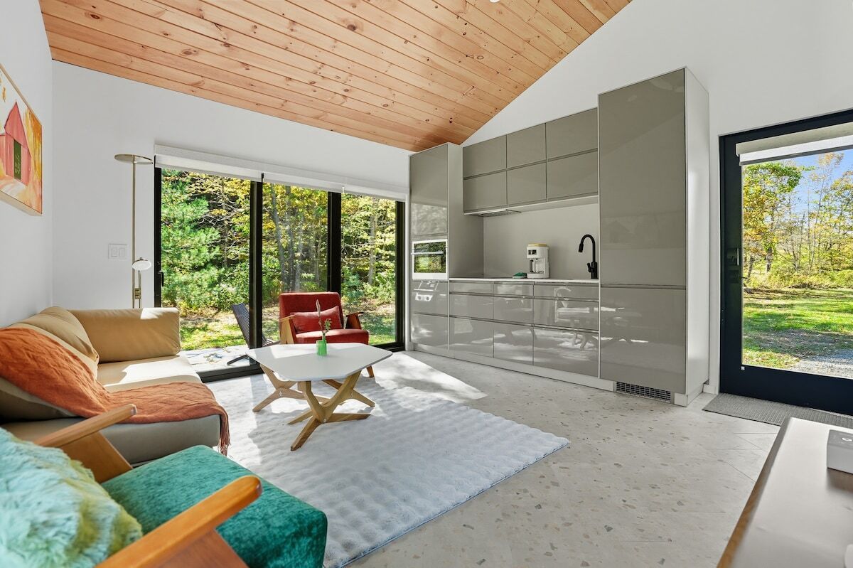 A modern living space with a sofa, a coffee table, and a red chair. There is a kitchen area with cabinets and a window.