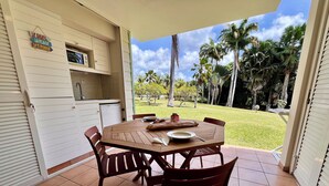 Een terras met een houten tafel en stoelen, een kitchenette met kastjes, en uitzicht op een gazon met palmbomen.