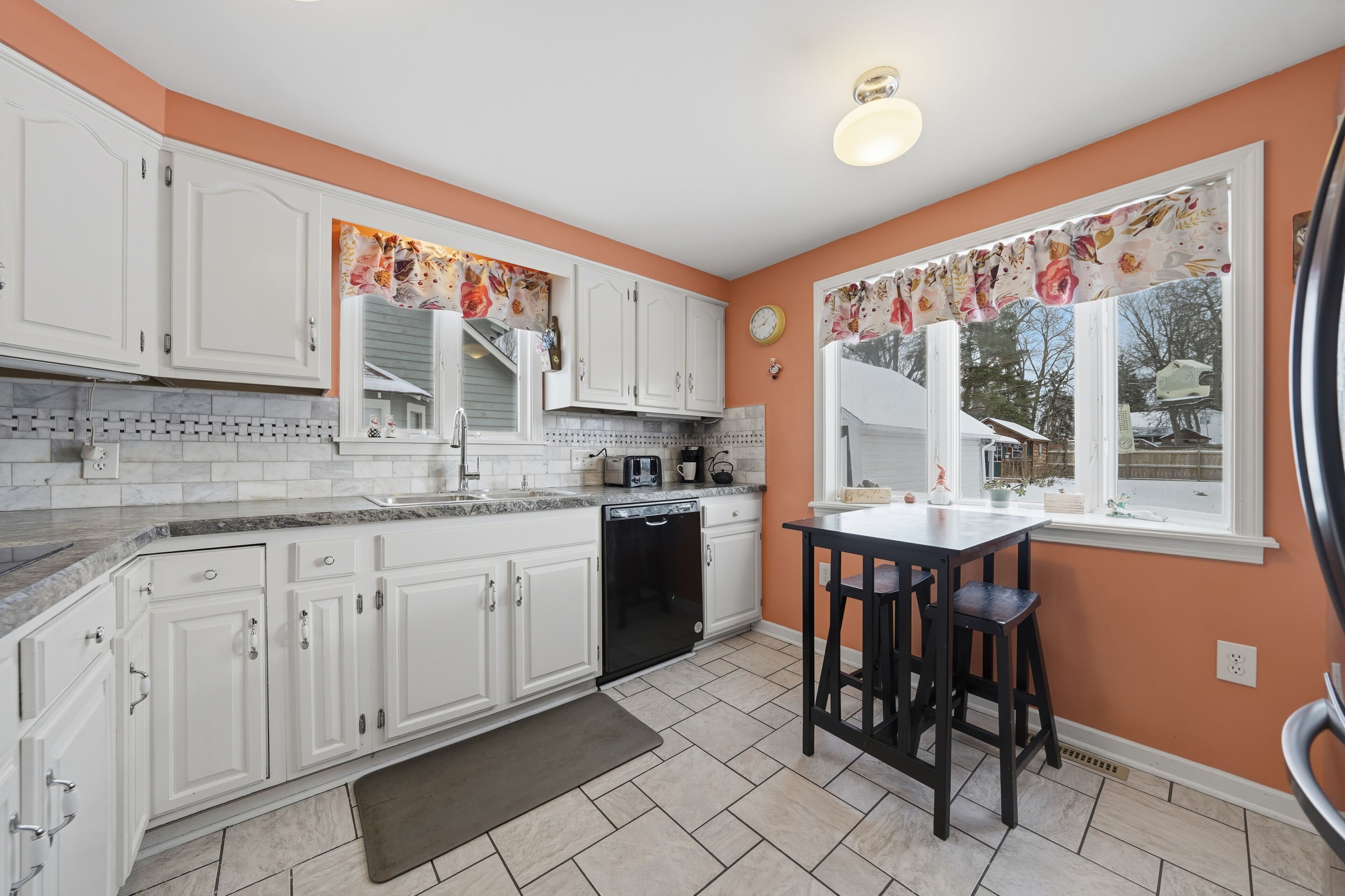 Una cocina con gabinetes blancos, un lavavajillas negro, una encimera con un tostador y una ventana con cortina de flores.