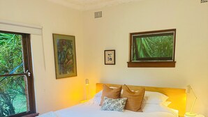 Un dormitorio con cama de madera, ropa de cama blanca, dos cuadros enmarcados, una lámpara de pie y una ventana con vista a la vegetación.