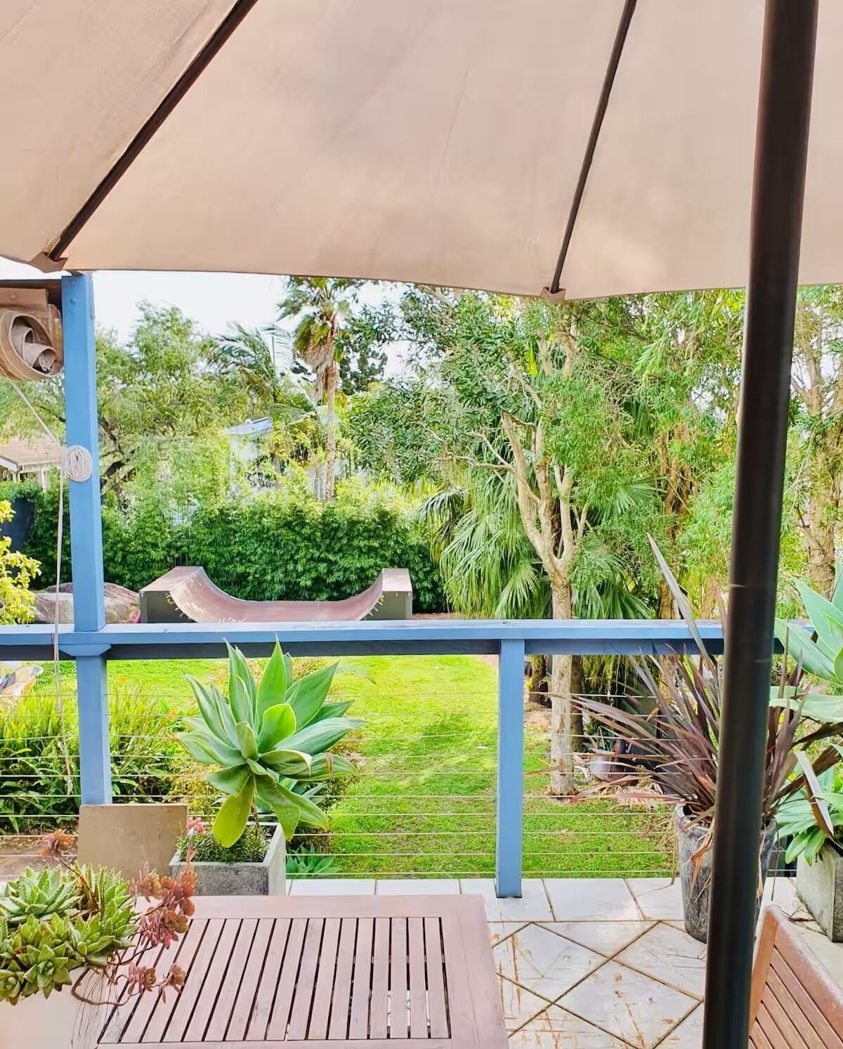 A covered patio area with a wooden table and chairs, surrounded by potted plants and a view of a lush garden.
