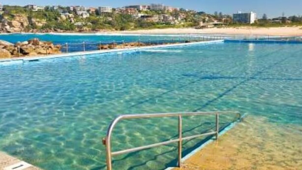 Una piscina cristalina con barandilla metálica, una playa al fondo y edificios en una ladera.