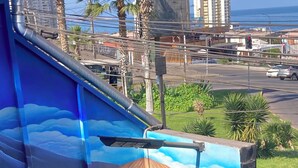 A wooden structure with a mural of a boat on water, a street view with cars, and palm trees in the background. - Spacious and Charming Apartment in Iquique. (Iquique)