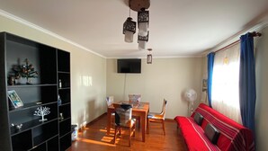 A living room with a wooden floor, a red sofa, a dining table with chairs, a black shelving unit, a TV mounted on the wall, and a window with blue curtains. - Spacious and Charming Apartment in Iquique. (Iquique)