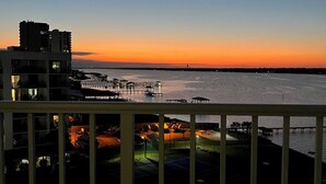Un balcon offrant une vue sur un coucher de soleil se reflétant sur une étendue d’eau, avec un quai et des bâtiments en arrière-plan.