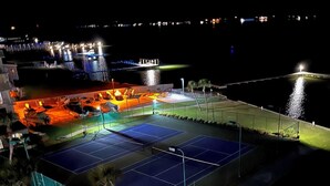 Un court de tennis extérieur entouré de sièges et doté d’un espace piscine, le tout éclairé par un système d’éclairage artificiel.