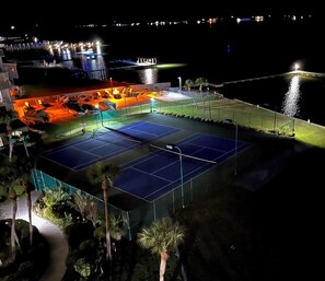 An outdoor tennis court with surrounding seating and a pool area, illuminated by artificial lighting. - Newly listed!  Beach, pool, hot tub, fishing, tennis, sunsets, and more! (Gulf Shores)
