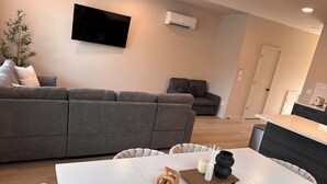 Un salon moderne avec un canapé gris, une table blanche surmontée d’un plateau en bois, et un coin cuisine à l’arrière-plan.